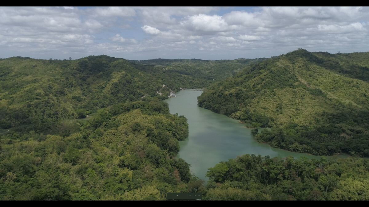 The image features an aerial shot of the Ipo Watershed on a cloudy day. Overlooking the mountains lush with greenery is a wide river in between hills connecting to the Ipo Dam in the distance.