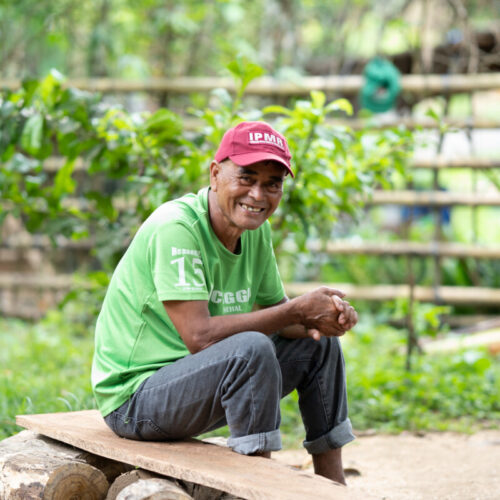 The image shows a 63-year old man sitting down on a piece of wood with his hands together. He is smiling while wearing a green shirt and a red cap against a backdrop of wooden fences and plants.